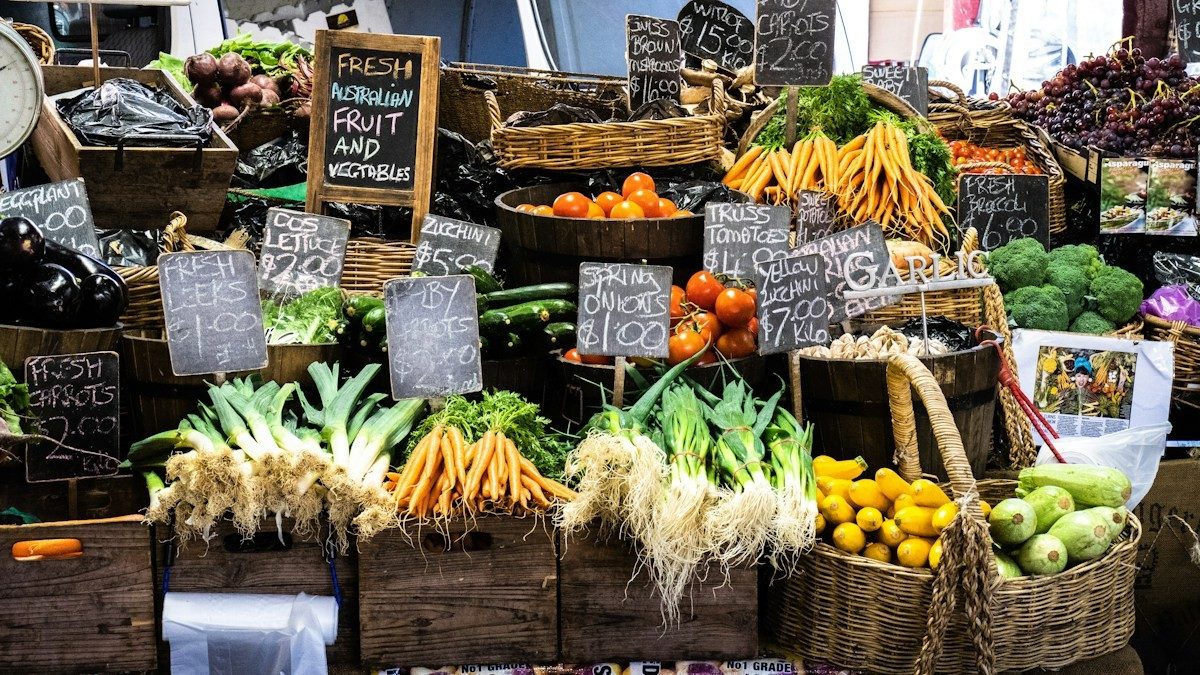Étal de marché couvert de caisses de légumes et de bananes, avec des ardoises de prix
